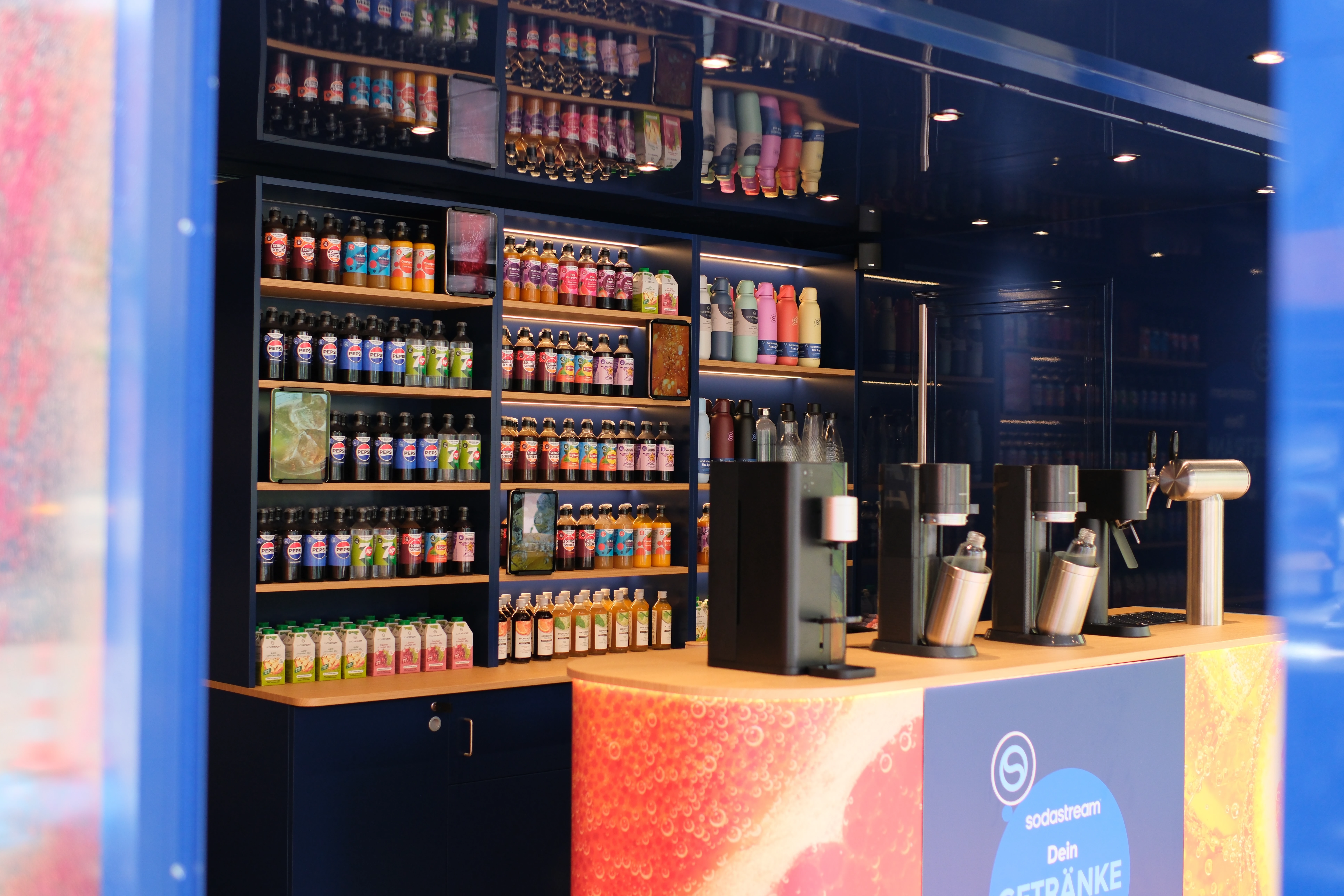 Moderne Getränkebar mit beleuchteten Regalen voller Flaschen und SodaStream-Geräten auf der Theke.